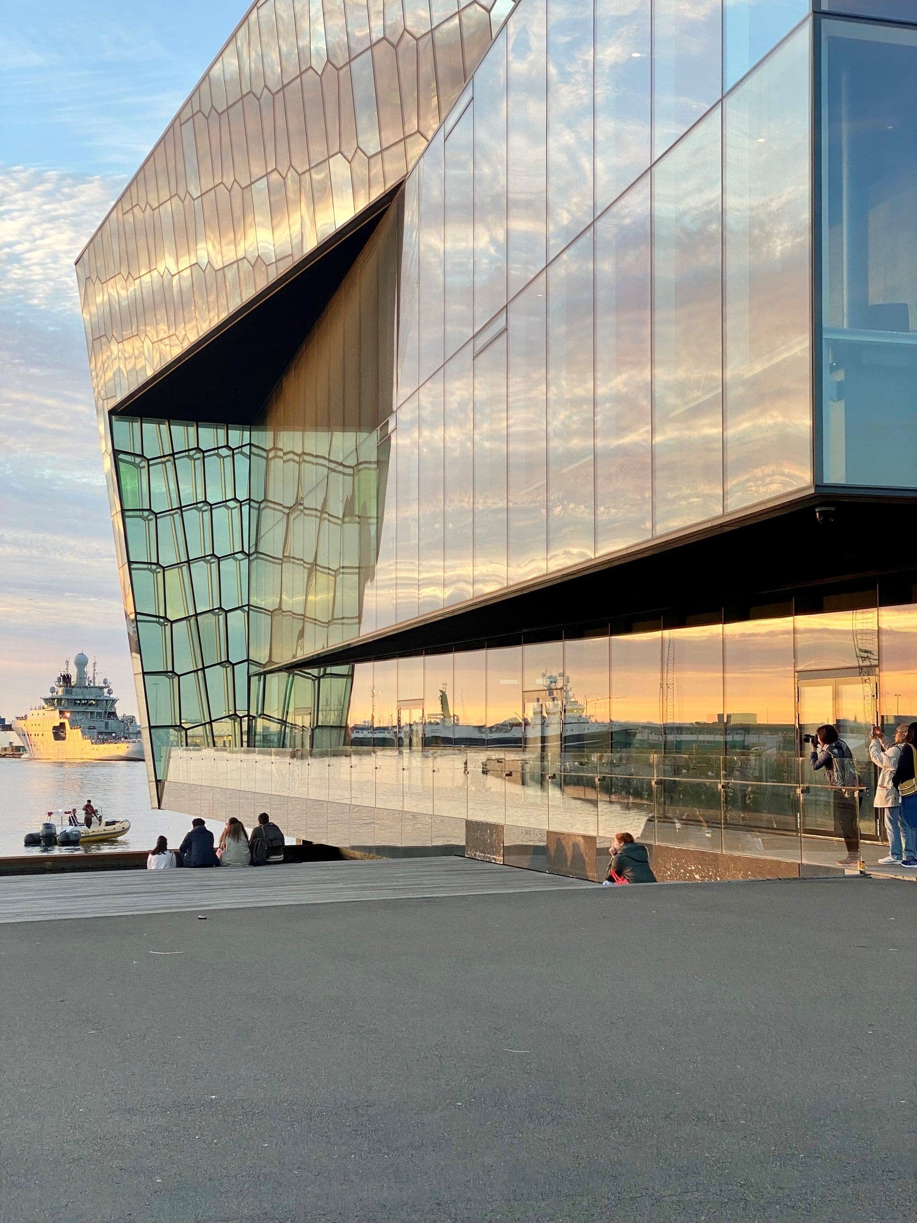 a group of people sit outside of a building with a boat in the background