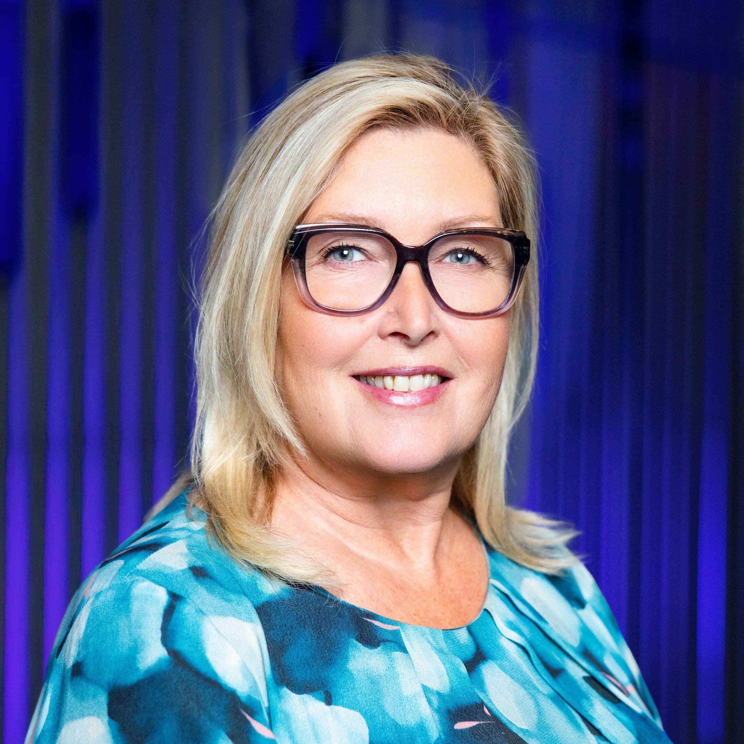 A smiling woman with blonde hair and glasses wears a blue patterned top against a blue background.