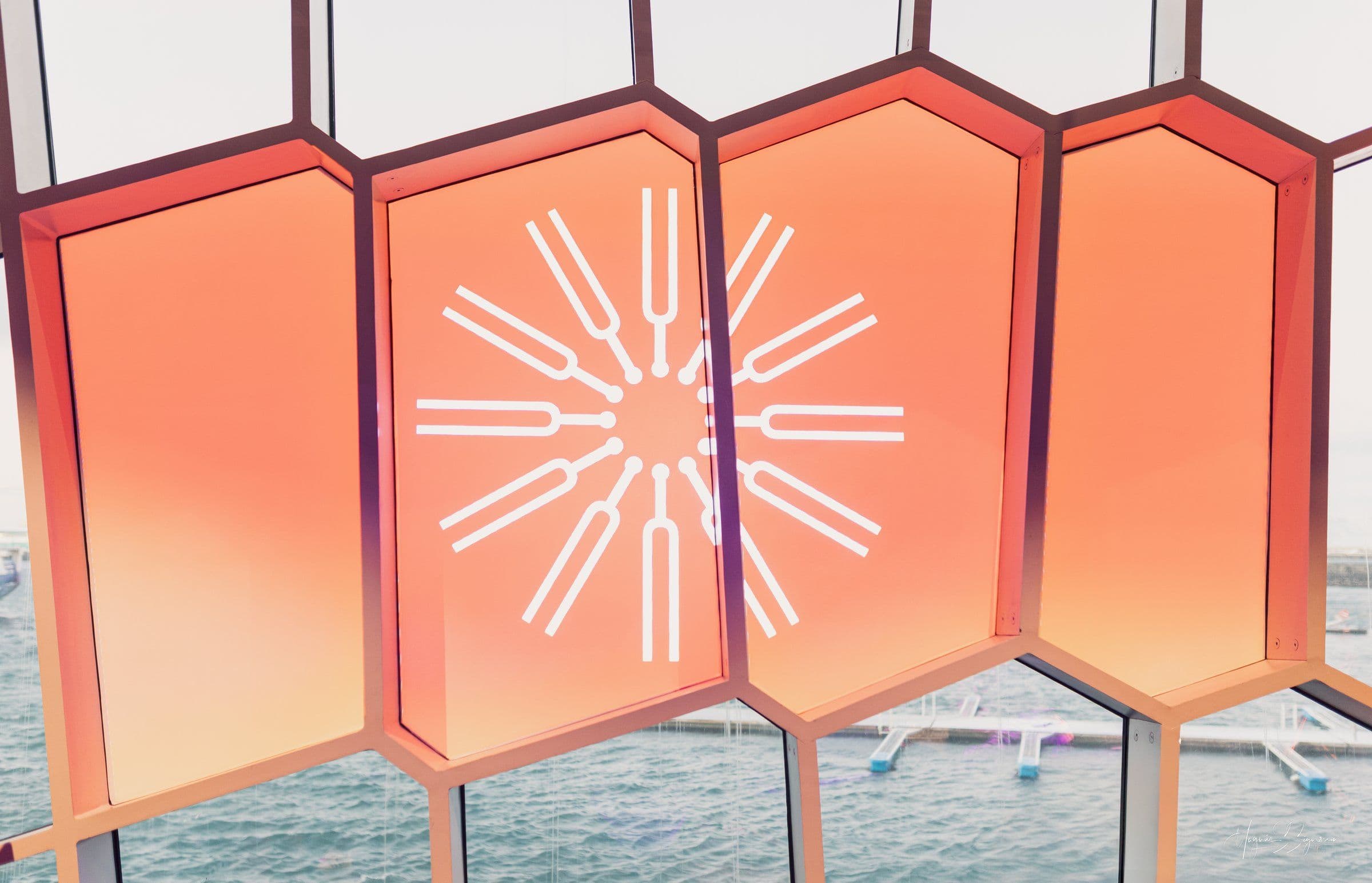 Orange hexagonal windows, one with a white circular tuning fork design, overlooking water.