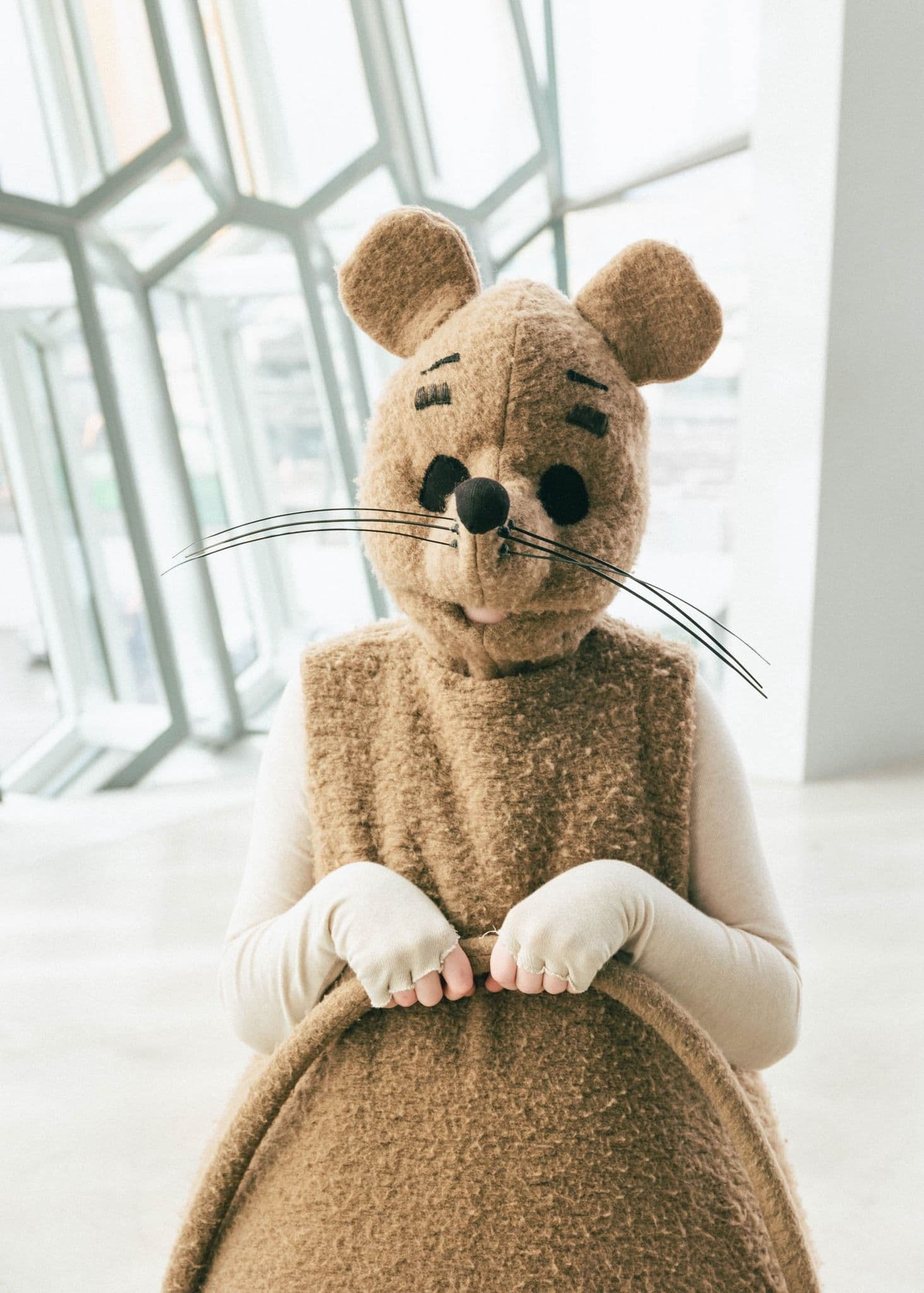 A person in a brown, fuzzy mouse costume with large ears, black eyes, nose, and whiskers stands indoors in front of modern geometric windows.